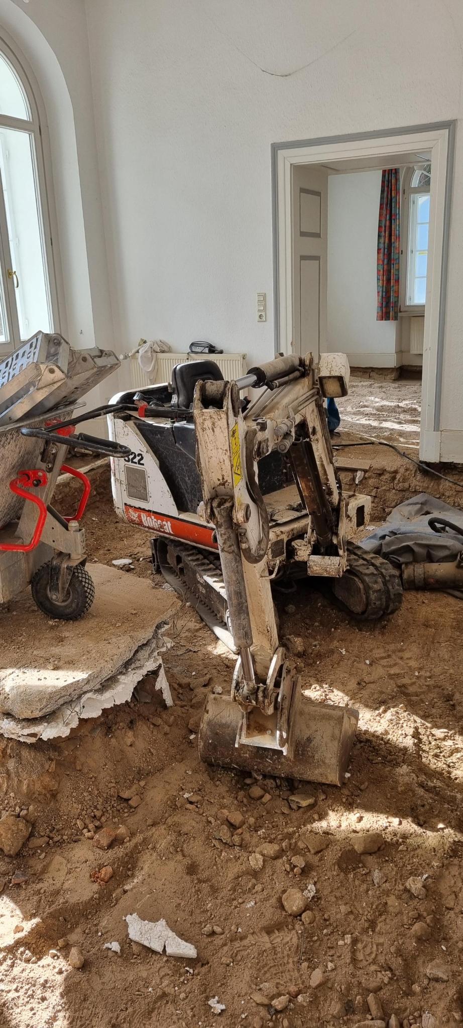 Ein kleiner Bobcat-Minibagger steht in einem entkernten, sandigen Innenraum während Renovierungsarbeiten.