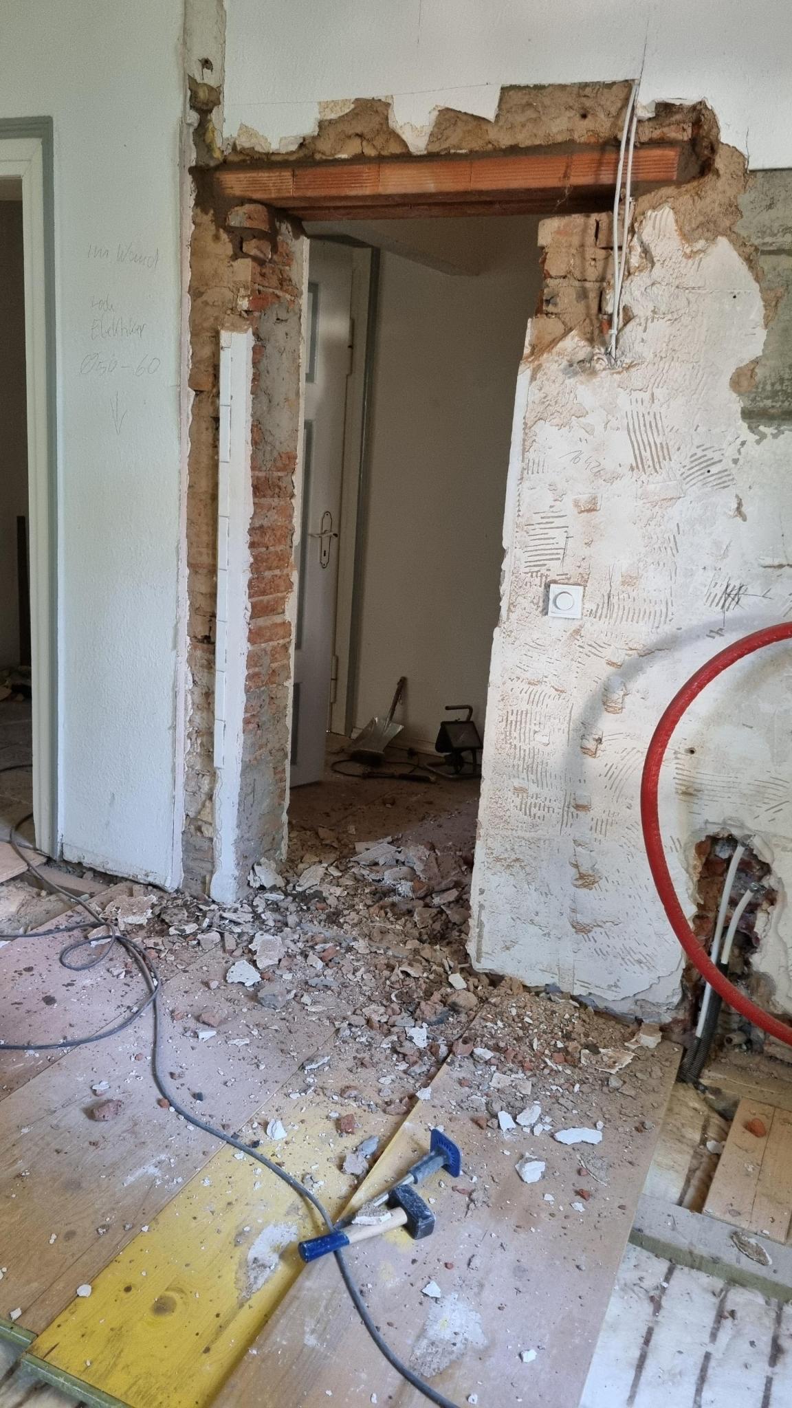 Ein unfertiger Wanddurchbruch in einem renovierungsbedürftigen Raum mit Bauschutt auf dem Boden und einem liegenden Hammer im Vordergrund.