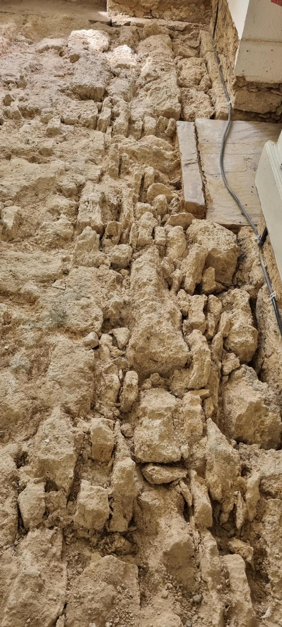 Nahaufnahme eines unebenen, rauen Untergrunds aus groben, hellbraunen Natursteinen neben einer hellen Hauswand.