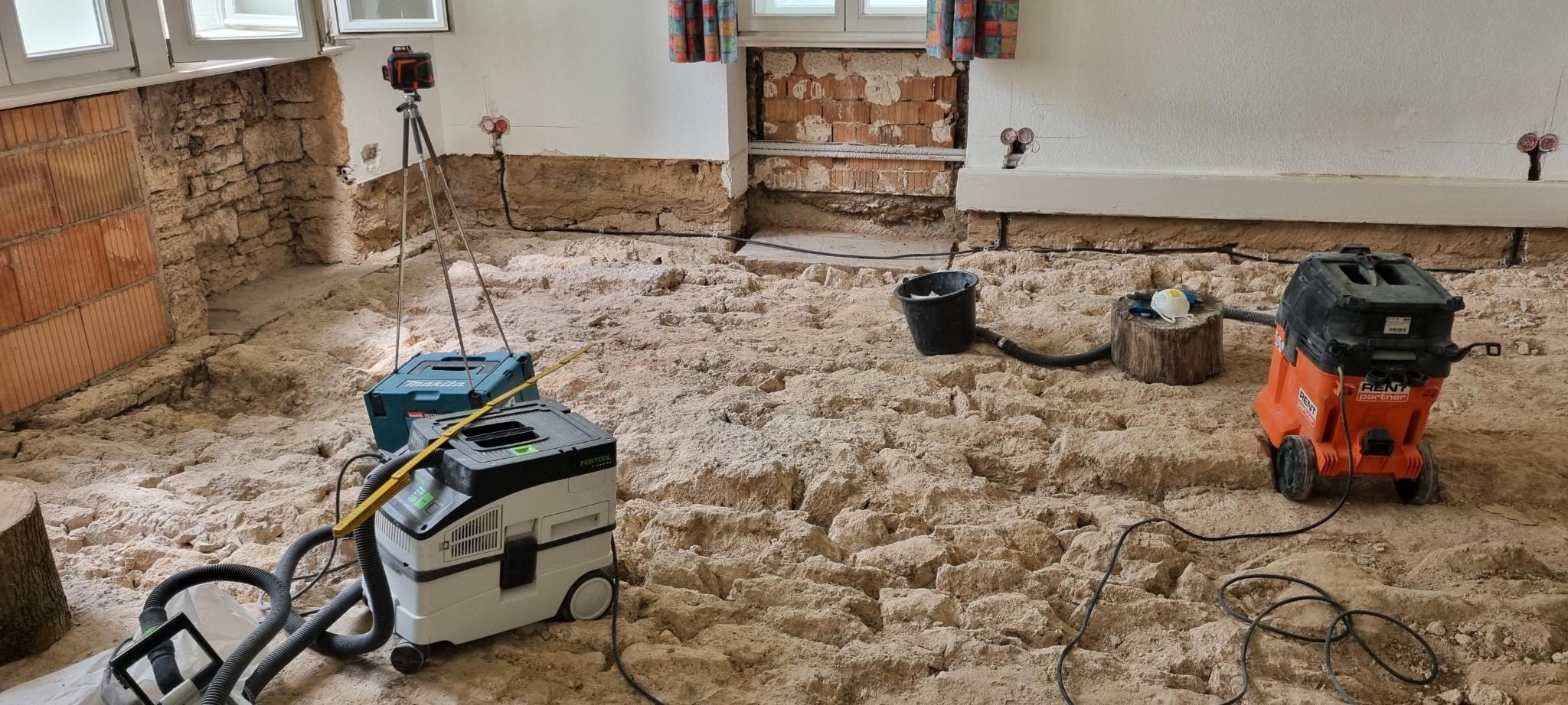 Ein Raum in der Sanierungsphase mit freigelegtem Untergrund, Baustellengeräten und einem Laser-Messgerät auf einem Stativ.